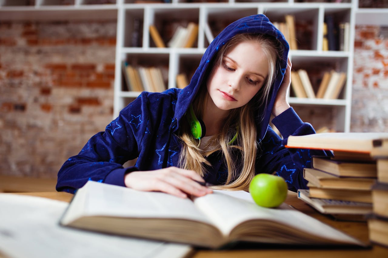 services-01 Teenage girl in blue hood reads in library, headphones and apple by her side.